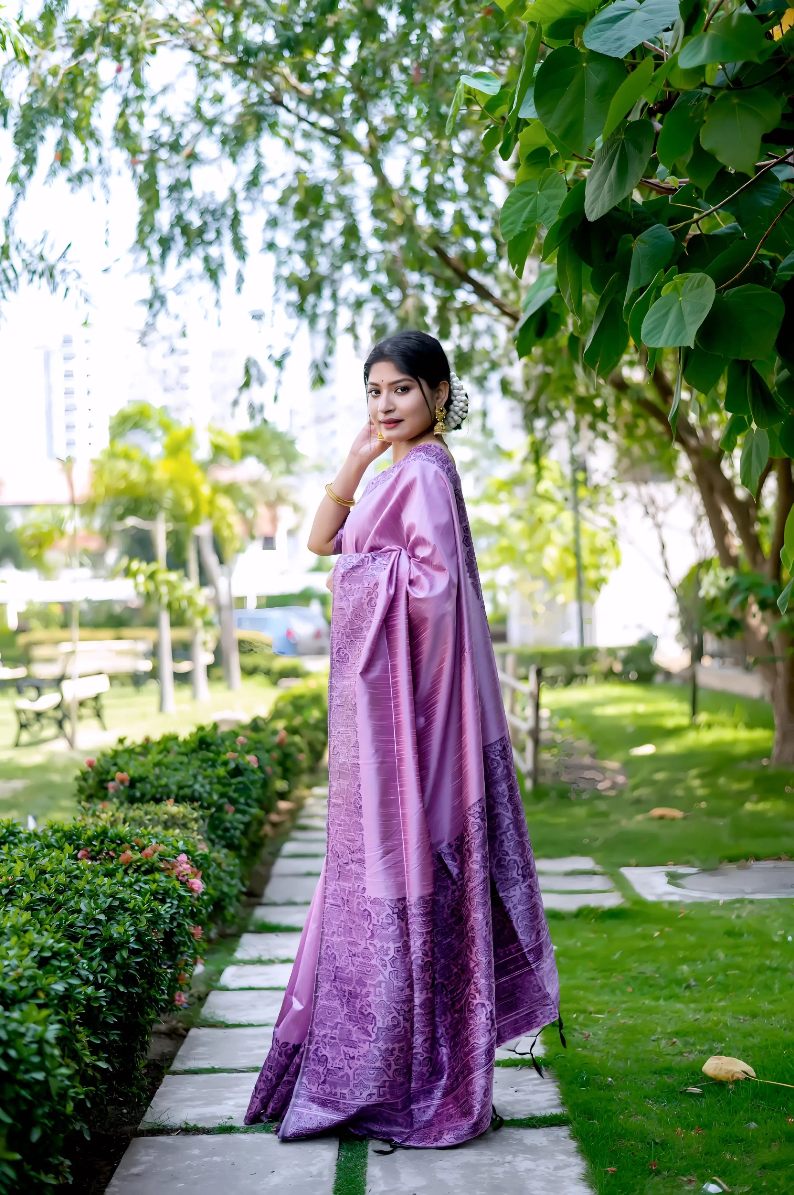 A woman models a luxurious Lavender/Violet Raw Silk Saree. The saree features a wide border and pallu with dense, tone-on-tone Kalamkari-style weaving in purple Zari. She wears a matching short-sleeve purple blouse and subtle gold jewelry, with flowers adorning her hair, posing outdoors on a stone pathway.